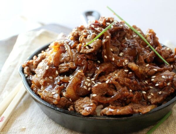 BEEF STIR-FRY HONEY SOY Sliced beef in glossy sauce with sesame seeds and green onions in a dark bowl.