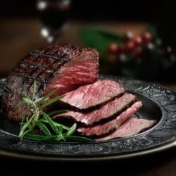 Sliced medium-rare grilled steak on ornate plate, garnished with rosemary sprigs.