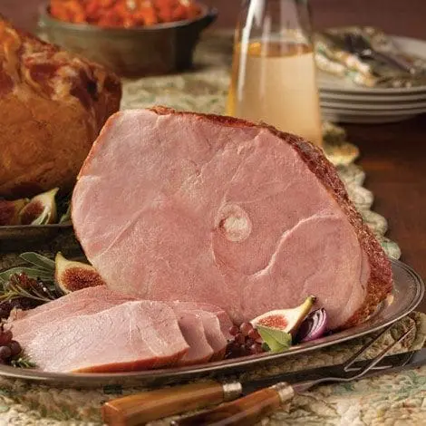 Cooked ham on decorative plate with slices, fruits, garnishes; background has dish and drink.