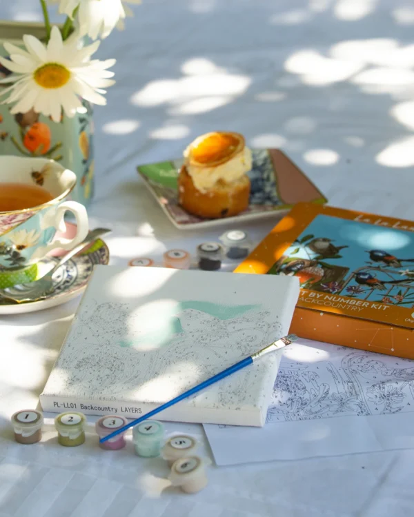 Outdoor paint-by-number kit setup with pastry, floral teacup, daisies, and sunlight on table.