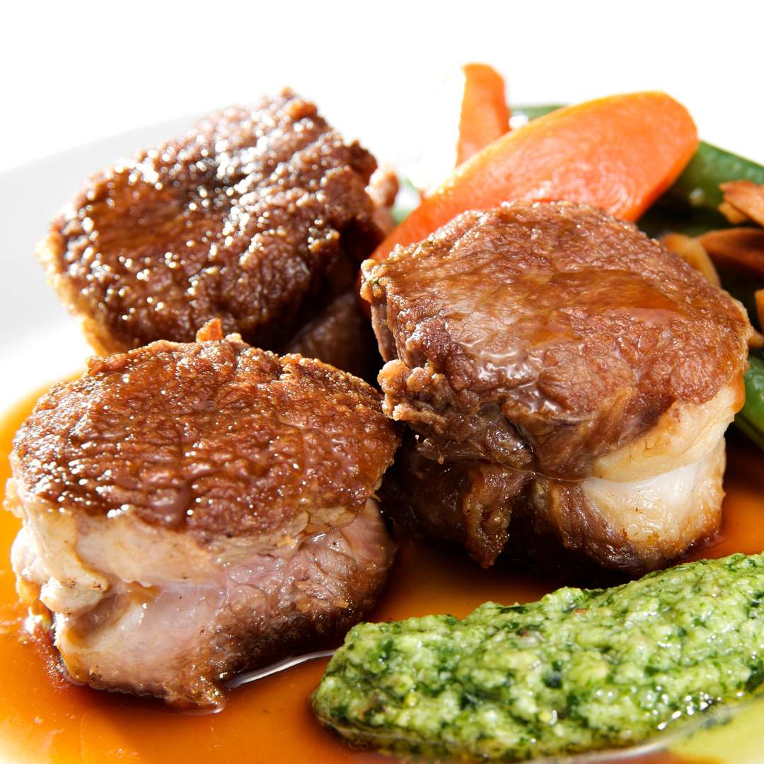 Cooked meat pieces in sauce with green puree, carrots, and green beans in the background.