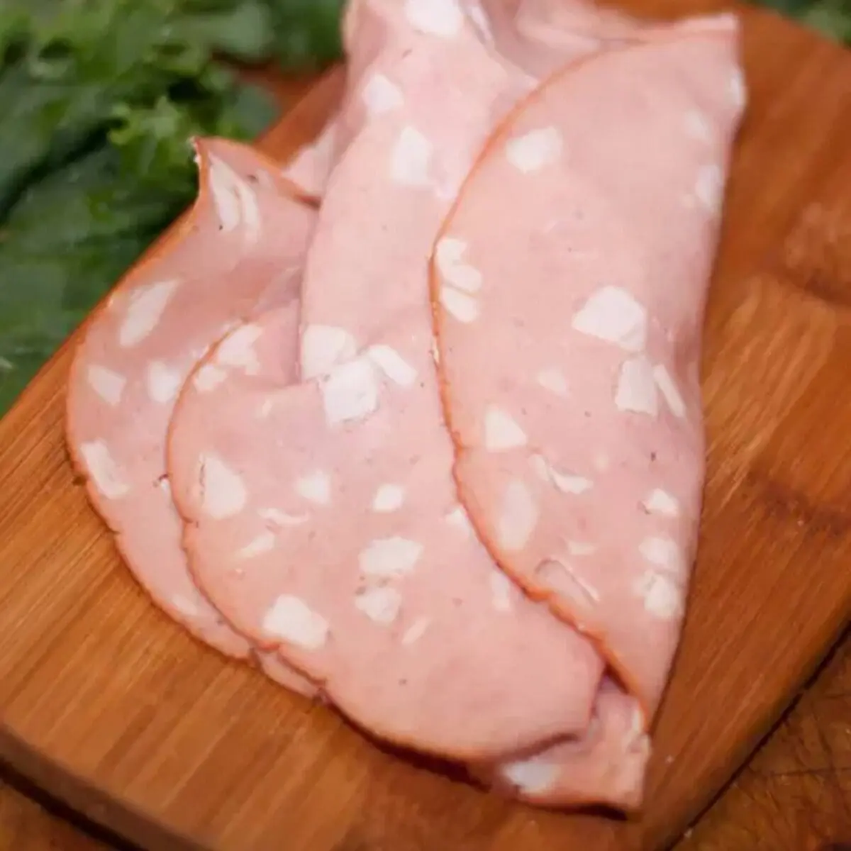 Thinly sliced mortadella with white specks on a wooden board, green herbs in background.