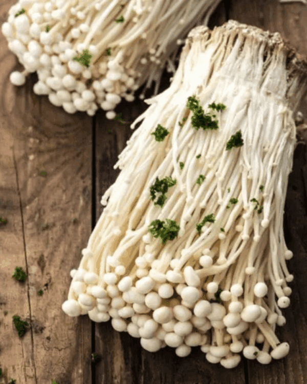MUSHROOMS FRESH ENOKI PACKETS Close-up of enoki mushrooms on wood, garnished with green herbs.