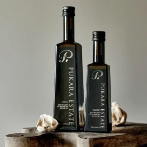 Two Pukara Estate garlic-flavored olive oil bottles on wood, surrounded by garlic cloves.