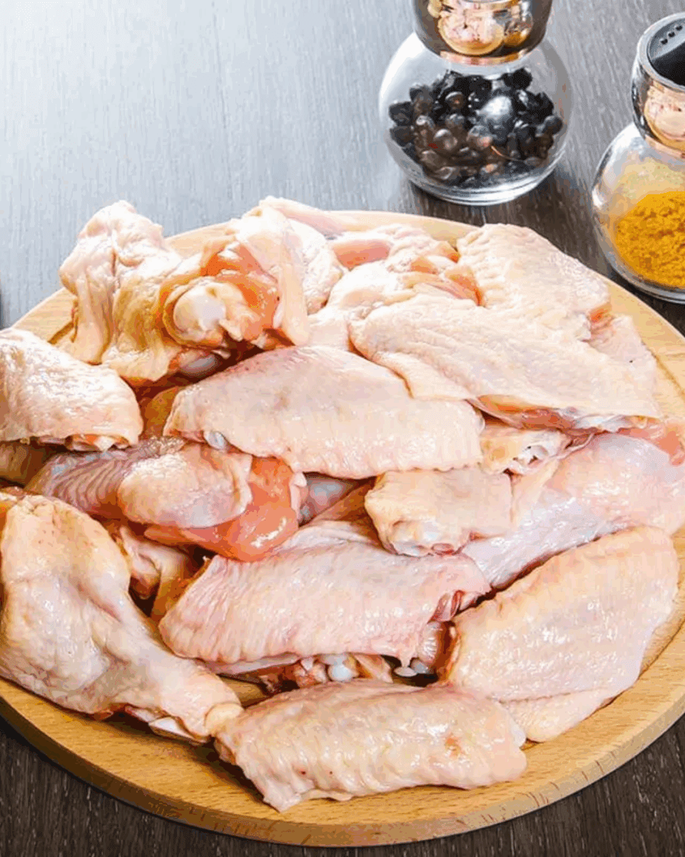 Wooden plate of raw chicken wings with spice containers on dark wooden surface.