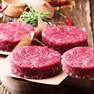 Raw burger patties on parchment paper with rosemary and chili peppers on a wooden board.