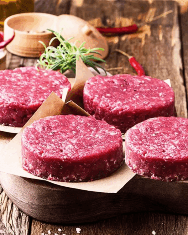 Raw burger patties on parchment paper with rosemary and chili peppers on a wooden board.