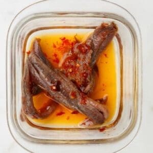 Glass container with anchovy fillets in oil, topped with red chili flakes.