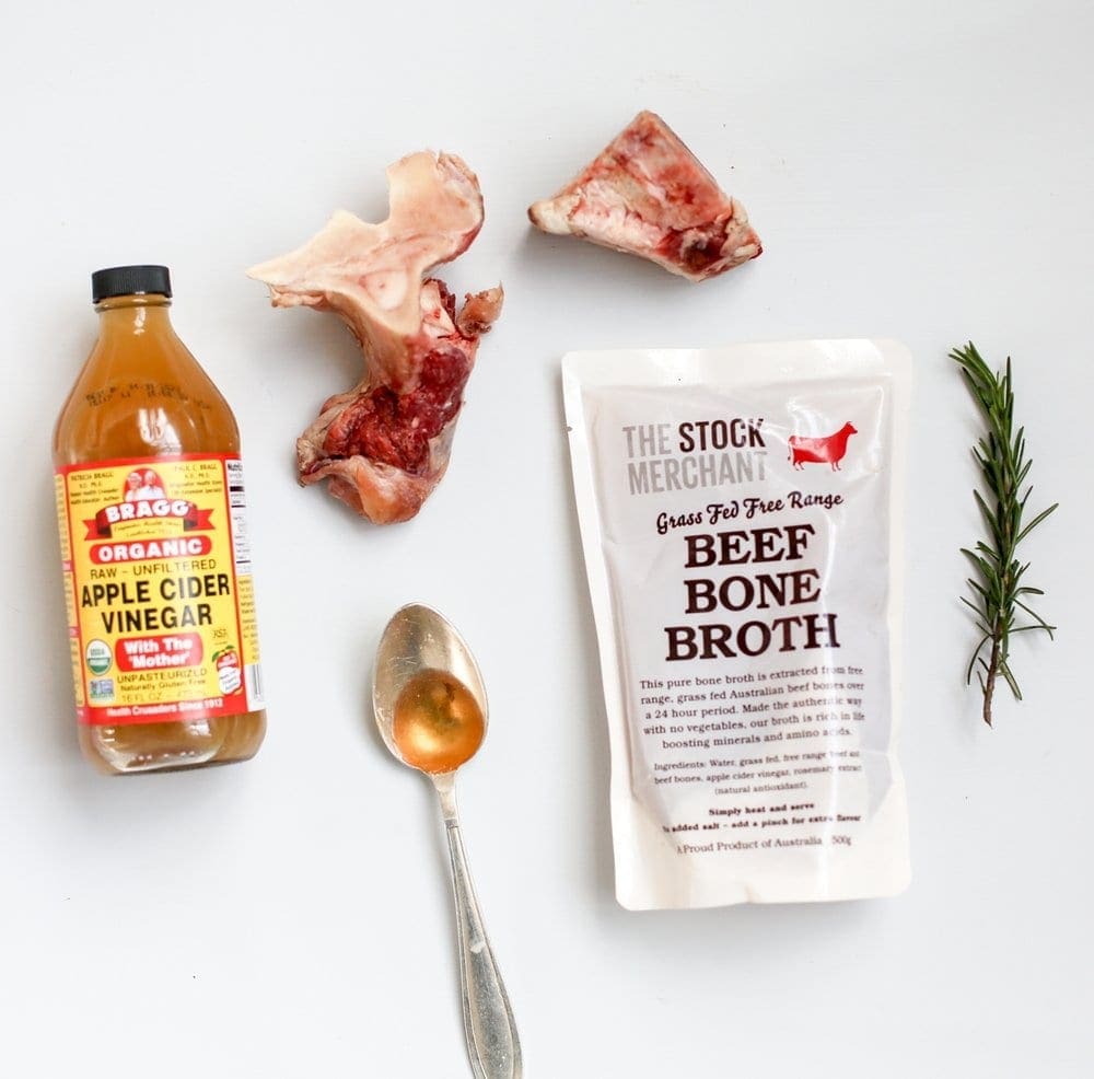 Apple cider vinegar, beef broth pack, beef bones, rosemary, and spoon on white background.
