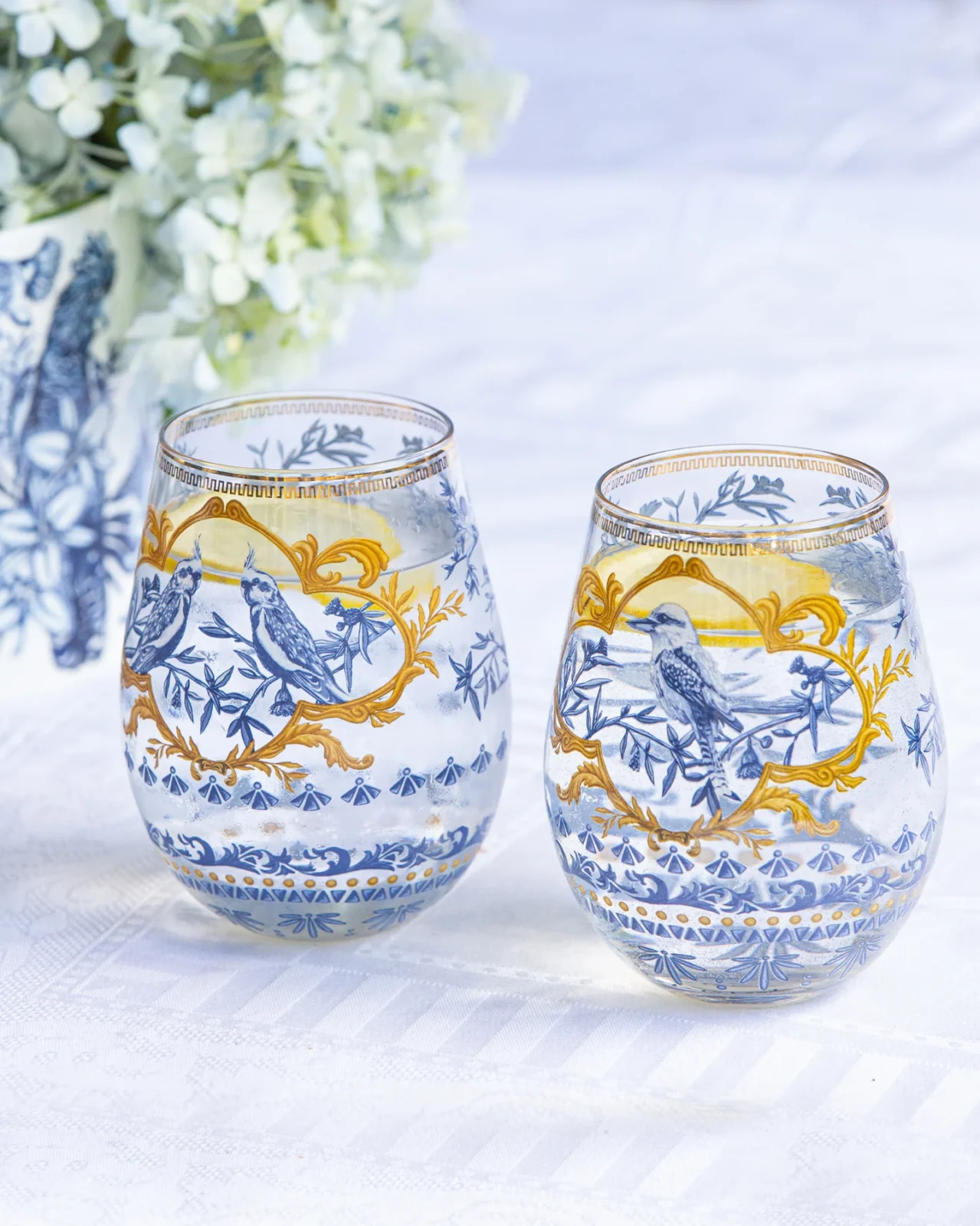 Decorative glass tumblers with blue bird and plant designs, gold accents, white flowers in background.
