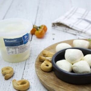 Bowl of bocconcini cheese with bread snacks, tub, tomatoes, and a towel on a wooden board.