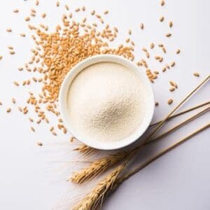 Bowl of flour surrounded by scattered wheat grains and stalks on a white surface.