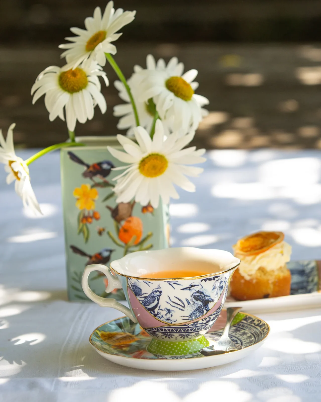 Ceramic teacup on saucer, vase with daisies, pastry on plate, sunny outdoor setting.