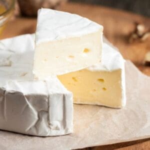 Round creamy cheese on parchment with a wedge cut out, walnuts, and honey on a wooden board.