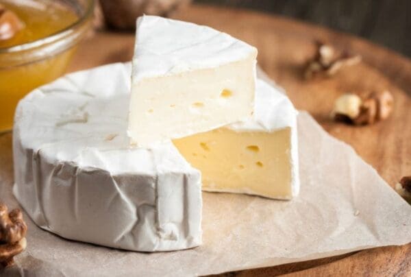 Creamy Cheese with Walnuts and Honey Round creamy cheese on parchment with a wedge cut out, walnuts, and honey on a wooden board.