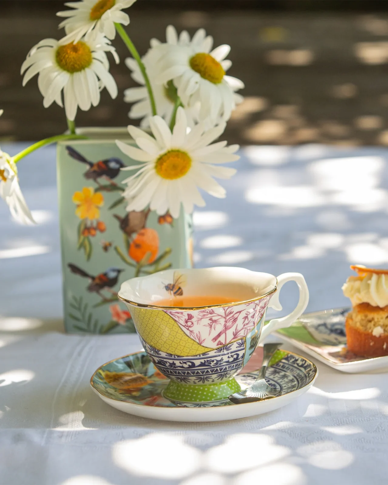 Decorative tea setup with teacup, saucer, cupcake, spoon, and vase of daisies on a tablecloth.