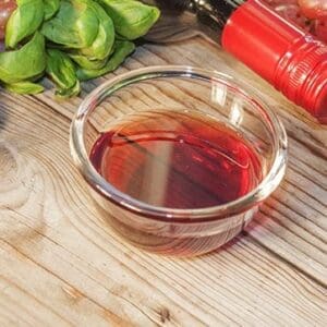Glass bowl with red liquid, basil, grapes, and bottle with red cap on wooden surface.