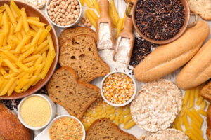 Overhead view of gluten-free foods including pasta, bread, black rice, corn, chickpeas, flour, and rice cakes spread across a wooden surface.
