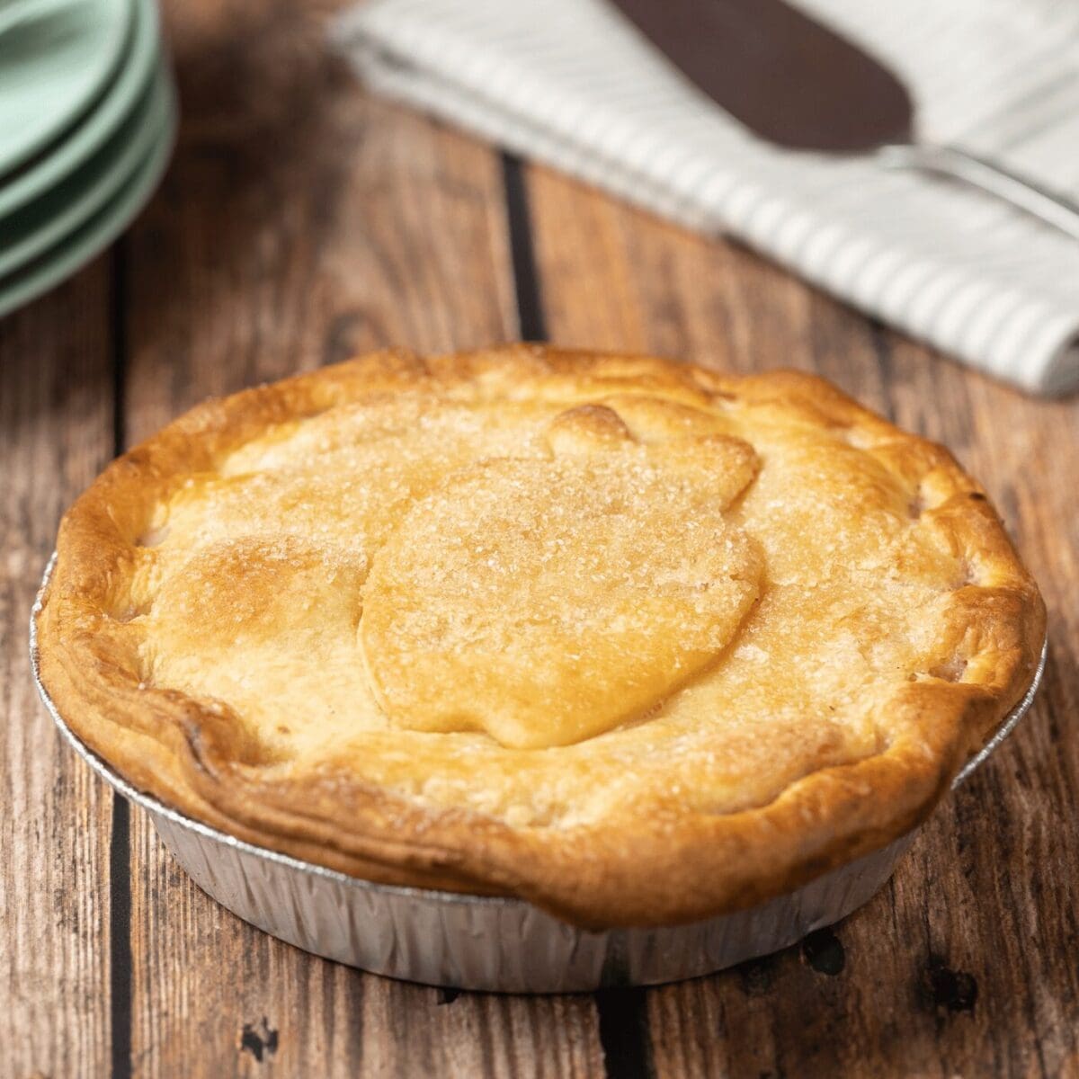 Golden-brown pie in tin with flaky crust, decorative top, green plates, and striped cloth nearby.