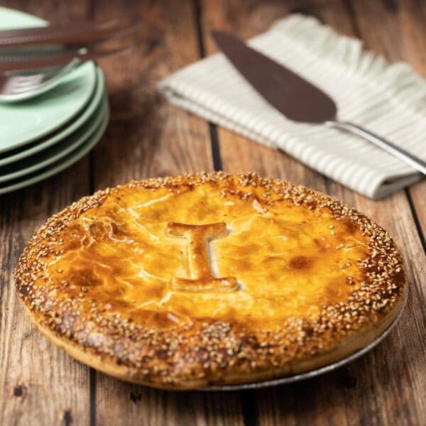 Golden-brown pie with sesame crust and embossed "I" on wooden table, plates, utensils nearby.