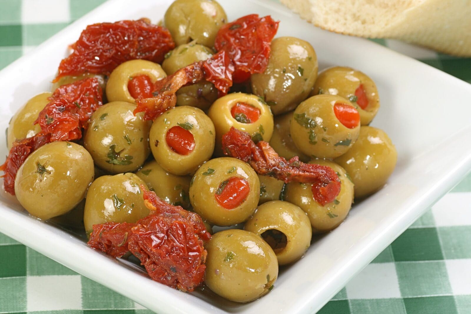 Green olives with pimentos, sundried tomatoes, herbs in white dish on checkered surface.