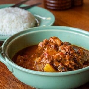Green bowl of stew with meat, vegetables on wooden table; plate of white rice beside.