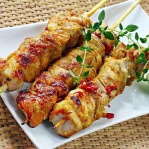 Grilled meat skewers with herbs and red pepper flakes on a white plate, wicker placemat background.
