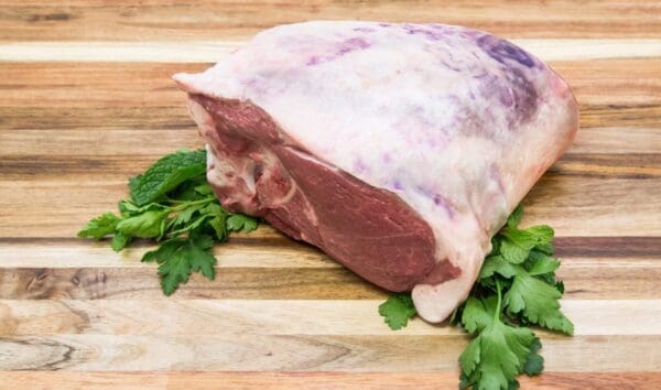 Raw Meat with Herbs on Cutting Board Large raw meat with fat on a wooden board, garnished with green herbs beneath.