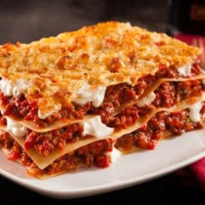 A plate of lasagna with layered pasta, meat sauce, cheese, on white plate, red napkin nearby.