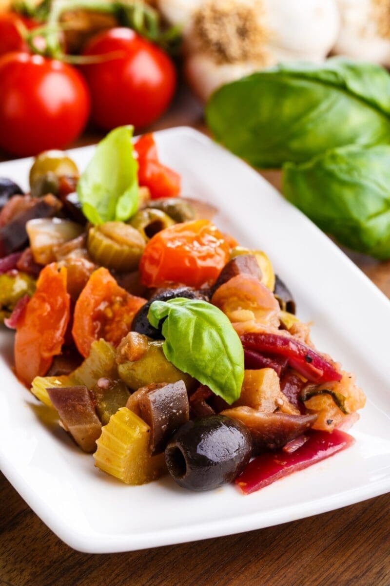 Plate of mixed vegetables with basil, including tomatoes, olives, and eggplant, on white plate.