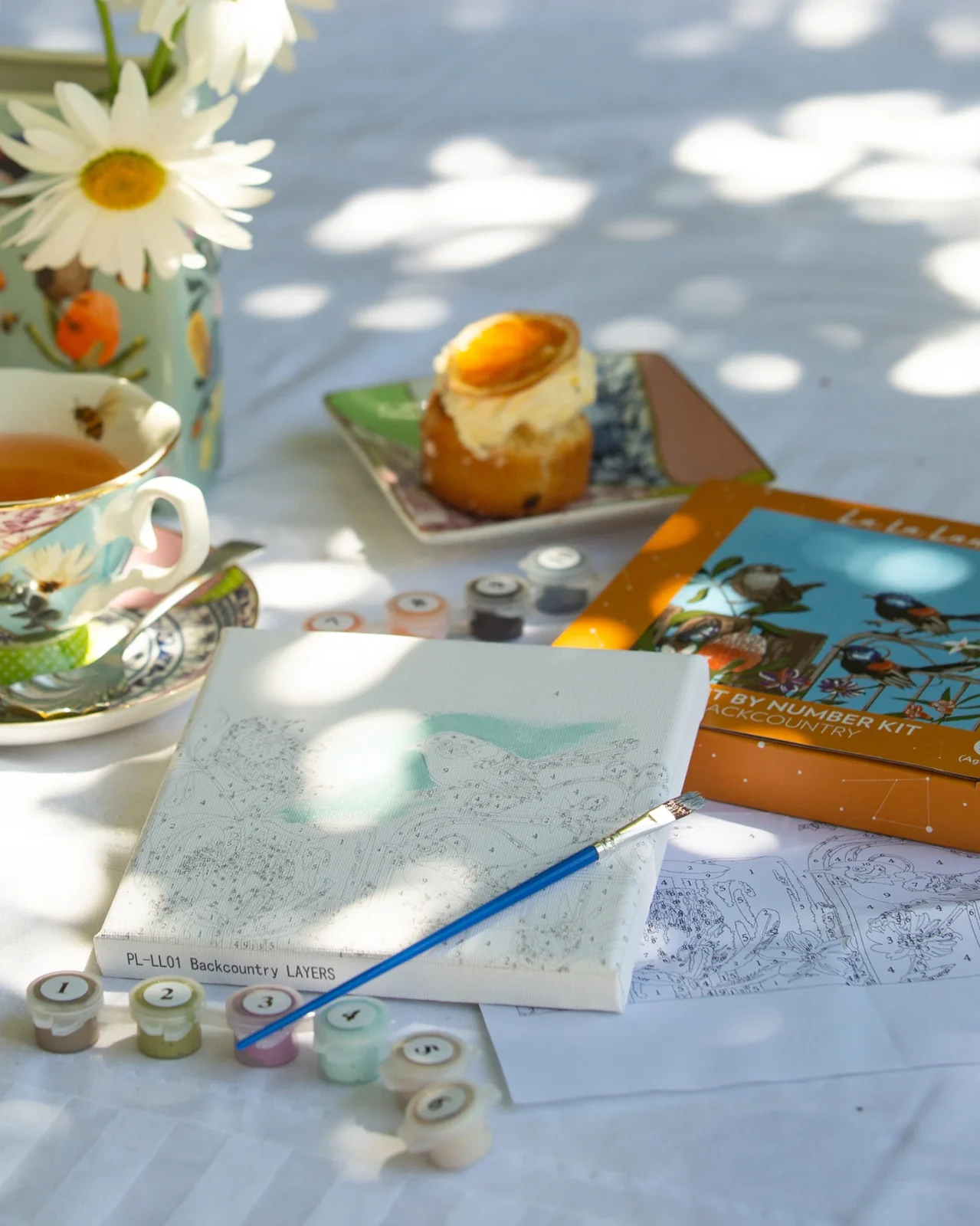 Outdoor paint-by-number kit setup with pastry, floral teacup, daisies, and sunlight on table.