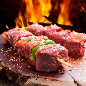Raw beef skewers with bell peppers, coarse salt, and flames on a wooden board.