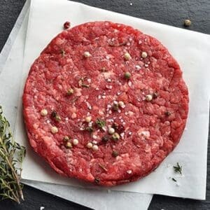 Raw hamburger patty on parchment with salt, pepper, herbs, and thyme on dark slate surface.