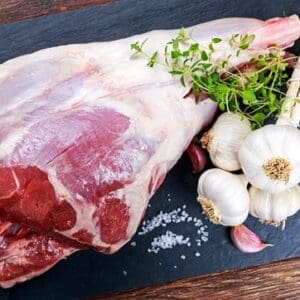 Raw lamb meat with garlic bulbs, cloves, fresh herbs, and coarse salt on a dark surface.