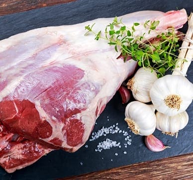 Raw lamb meat with garlic bulbs, cloves, fresh herbs, and coarse salt on a dark surface.