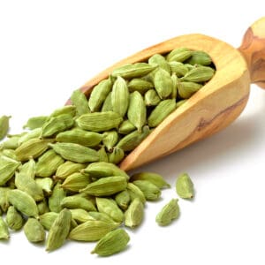 Wooden spoon with green cardamom pods on scattered white surface.