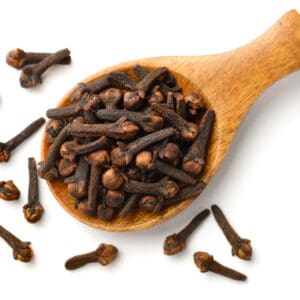 Wooden spoon filled with dried cloves on rustic wooden surface, scattered cloves nearby.