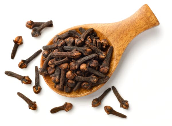 Wooden spoon filled with dried cloves on rustic wooden surface, scattered cloves nearby.