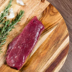 Raw steak on wooden board with rosemary and garlic clove nearby.