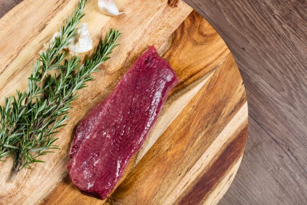 LAMB BACKSTRAPS Raw steak on wooden board with rosemary and garlic clove nearby.