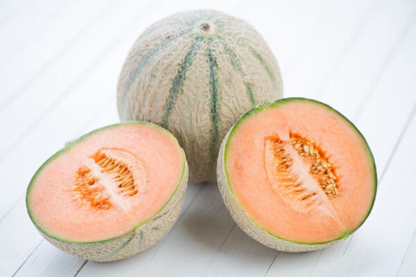 Whole cantaloupe and two halved cantaloupes on a white wooden surface, showing orange flesh.