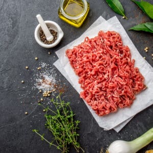 Raw ground meat on parchment paper with herbs, pepper, olive oil, salt, and bay leaves.