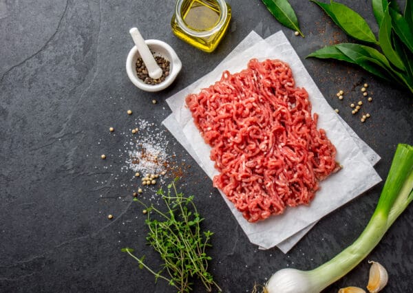LAMB MINCE Raw ground meat on parchment paper with herbs, pepper, olive oil, salt, and bay leaves.