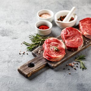 Raw beef steaks on wooden board with rosemary, spices, mortar and pestle, gray background.