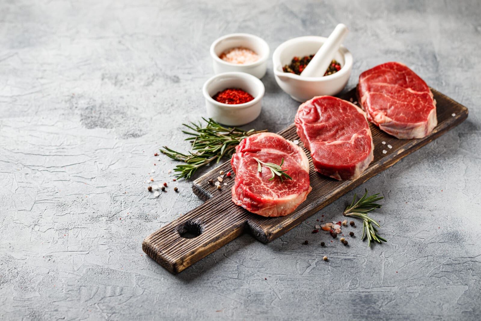 Raw beef steaks on wooden board with rosemary, spices, mortar and pestle, gray background.