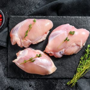 Two raw chicken thighs on a wooden board with a sprig of fresh herbs.
