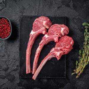 Three raw lamb chops, red peppercorns, and thyme on a black slate surface.