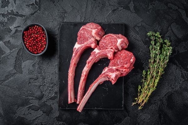 Lamb Chops with Fresh Mint Three raw lamb chops, red peppercorns, and thyme on a black slate surface.