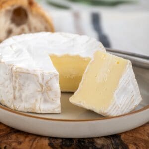 Soft cheese wheel with white rind, sliced to show creamy inside, on checkered cloth with honey.