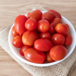 White bowl with small red tomatoes on burlap and wooden surface.